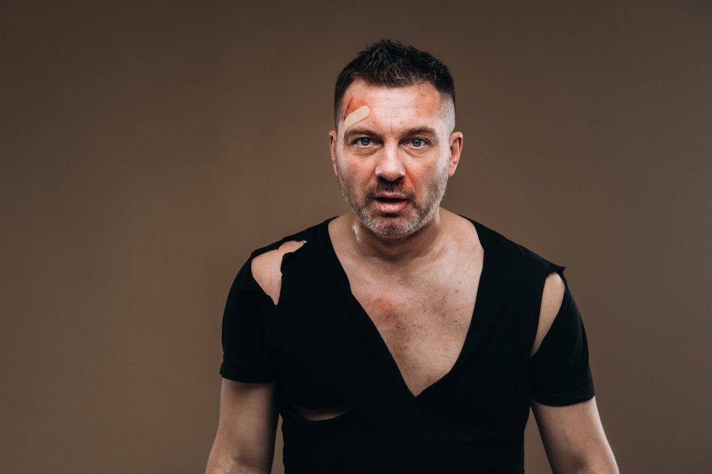 Against a gray background stands a battered angry man in a black T-shirt with wounds